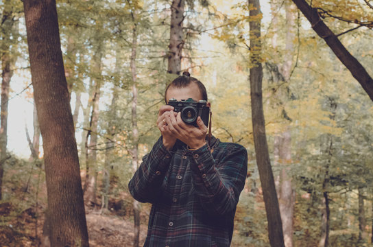 Hipster millennial young man or teenager with analog vintage photo camera makes photograph into lens in middle of old wood with sunset light, explores tourism. Hipster Travel Lifestyle concept