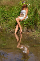 Pretty girl young woman with natural blond long hair and beautiful face in white dress and flower wreath sits on river beach on sunny summer day with cute brown rabbit