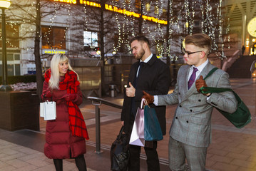 The happy, elegant young friends standing on the street and holding the shopping bags in hands and showing thumbs up