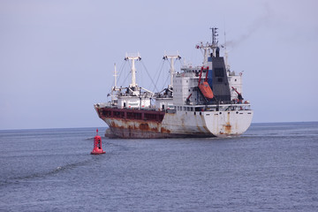A rusty reefer ship leaving harbor next to a port buoy marker 