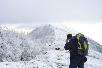 Naklejka na ściany i meble Hiker with backpack and camera taking picture of snowy mountain. Winter mountain landscape, Happy tourist hiking in winter 
