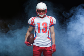 Fototapeta premium Front view portrait of american football player wearing helmet with ball against white smoke
