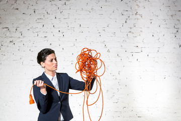 Confident young businesswoman looking at tangled red cable against white brick wall