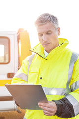 Confident tow truck driver making reports on clipboard against sky