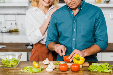 cropped image of mature wife hugging husband while he cooking salad in kitchen