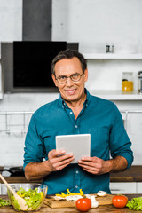 smiling handsome mature man using tablet while cooking in kitchen, looking at camera