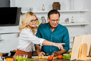 mature wife pointing on cookbook to husband in kitchen