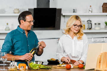 mature wife cutting vegetables and husband holding bottle of wine in kitchen