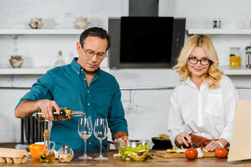 handsome mature husband pouring wine in glasses, attractive wife cutting vegetables in kitchen