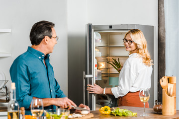 mature wife taking asparagus from fridge and husband cutting vegetables in kitchen