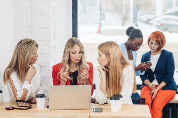 Group of young women co-workers discussing creative project during work process, sitting at office with laptop and take-away coffee. Blonde business woman explains main ideas to her colleagues.