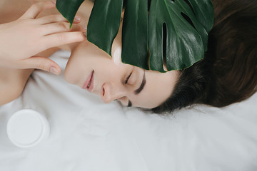 Portrait of a young long-haired girl applying cream on her face lying on the bed with a green leaf...