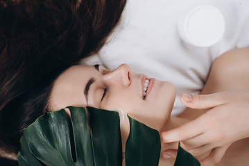 Fototapeta premium Portrait of a young long-haired girl applying cream on her face lying on the bed with a green leaf enjoying and relaxing . Lovely brunette with attractive appearance. Skin Care spa relax concept.