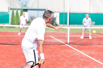 Man swinging racket while playing tennis doubles on red court during summer weekend