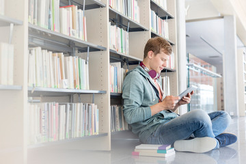 Student using digital tablet against bookshelf while sitting on floor at library - obrazy, fototapety, plakaty