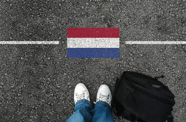  a man with a shoes and backpack is standing on asphalt next to flag of Netherlands and border