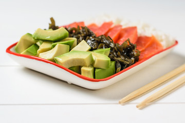 Bowl with rice, seaweed, avocado slices and fish on a white table with wooden sticks.
