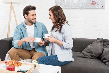 Obraz premium smiling couple sitting on couch and drinking coffee during breakfast at home
