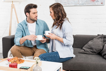 couple sitting on couch, looking at each other and drinking coffee during breakfast at home