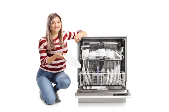 Beautiful young woman putting a plate into a dishwasher and smiling at the camera