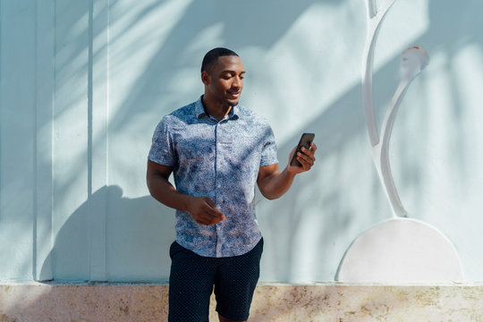 Smiling Young Man Wearing Shirt Looking At Cell Phone At A Wall