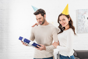beautiful smiling couple with gift box embracing and celebrating birthday in living room