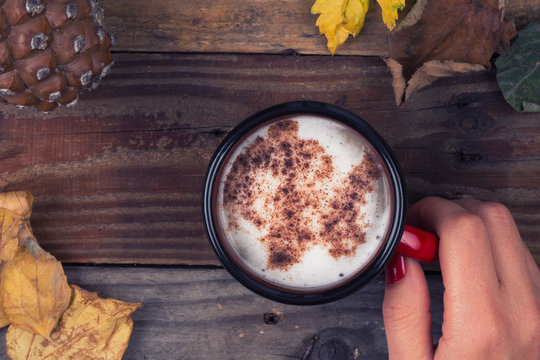 Woman hand holding coffee on wooden table. Dry leaves and a cone around.