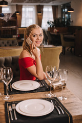 attractive woman in red dress sitting in restaurant using smartphone and looking at camera