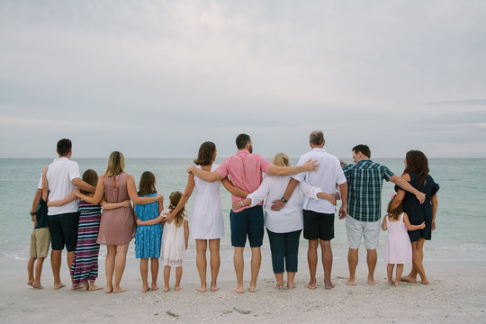 Huge Happy Traveling Caucasian Family with Adults and Kids at the Beach Hugging and Embracing while facing the Ocean Horizon Outside on Destination Vacation