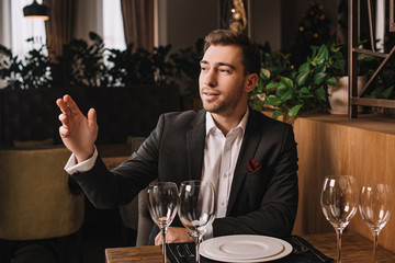 handsome man in suit waiting for girlfriend in restaurant and rising hand up