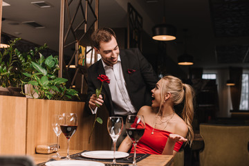 handsome boyfriend giving red rose to surprised girlfriend in restaurant