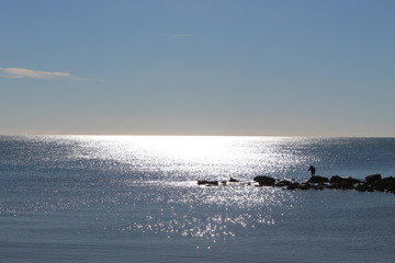 Controluce di pescatore sugli scogli con sole riflesso nel mare e cielo azzurro
