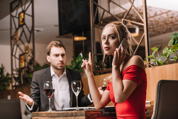 dissatisfied man looking at girlfriend talking on smartphone during dinner