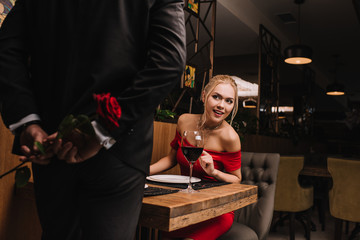 curious girl looking at man hiding red rose in restaurant