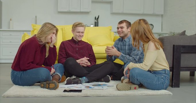 Positive smiling diverse college students sitting on the floor in loft apartment and communicating while taking a break from study. Cheerful carefree group of students relaxing after college at home.