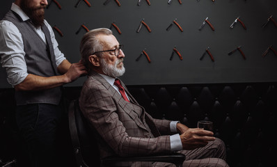 Professional male barber cutting hair of grey- haired wealthy man in years, dressed in fashionable...