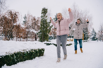 joyful woman running from boyfriend throwing snowball in winter