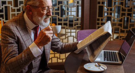Indoor portrait of a confident and handsome business mature European man wearing spectacles, looking at newspaper laptop holding a cup of coffee while sitting at a table with laptop in a cafe