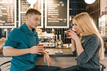 Young smiling man and woman together talking in coffee shop sitting near bar counter, couple of friends drinking tea, coffee