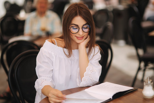 Stylish lady sits in cafe in centre of city and look at the menu. Sunny weather and codl drink. Girl in sunglasses chooses what to order