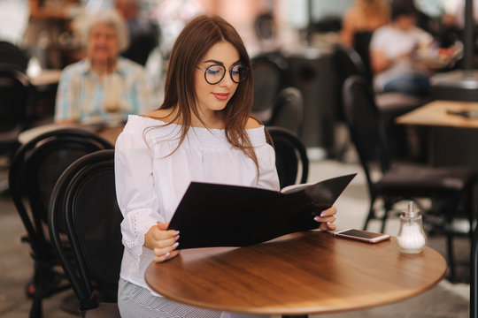Stylish lady sits in cafe in centre of city and look at the menu. Sunny weather and codl drink. Girl in sunglasses chooses what to order