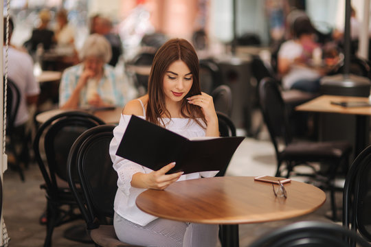 Stylish lady sits in cafe in centre of city and look at the menu. Sunny weather and codl drink. Girl in sunglasses chooses what to order