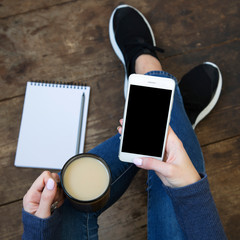 The girl sits on a wooden floor and holds a smartphone with a blank screen and latte in her hands....