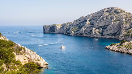 Fototapeta na wymiar Panoramic view of the cap Morgiou on the mediterranean shore near Marseille, France, with motorboats cruising and sailboats mooring in the blue waters of the calanque de Morgiou on a sunny day.