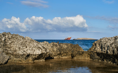 A red cargo ship wreck in the distance stranded on a small island. Rocky seashore landscape in Greece with blue sky and vivid clouds in summer.