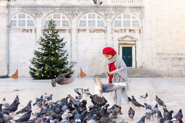 Kid girl is feeding doves on square near Christmas tree in Europe. Pigeons are flying around cute...