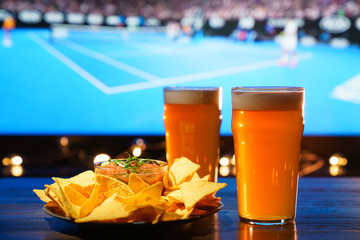 2 glasses of beer with nachos and salsa on a dark wooden table. Sports bar, tennis on a background,...