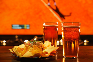 2 glasses of beer with nachos and salsa on a dark wooden table. Sports bar, tennis on a background,...