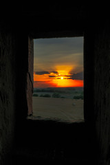 Sonnenuntergang durch ein Burgfenster Fotografiert.Im Vordergrund Feld und Wiesen die Sonne steht in der Mitte vom Bild.