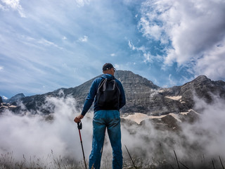 A man stands on a slope and admires the tops of mountains in the clouds.